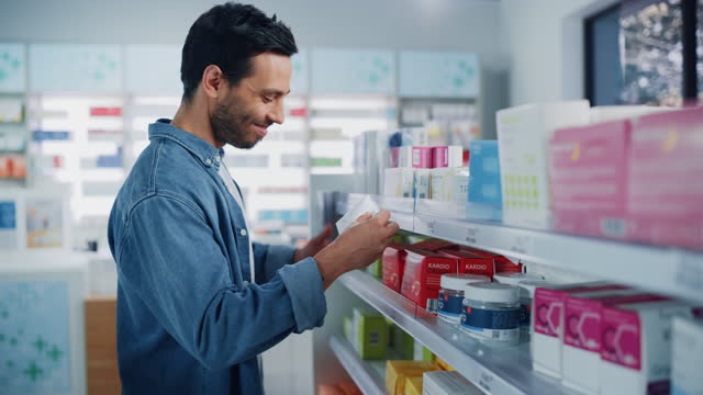 Pharmacy Drugstore: Portrait of Handsome Latin Man Choosing to Buy Medicine Browsing through the Shelf, Successfully finds what he Needs, Smiles Happily. Modern Pharma Store Health Care Products