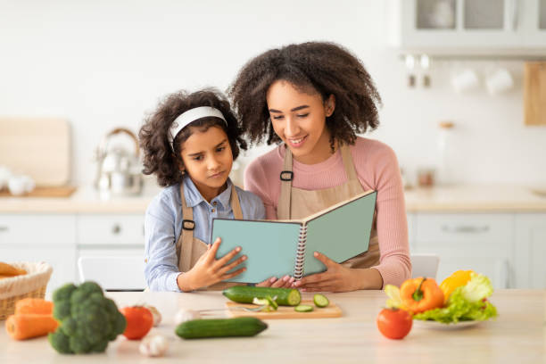 femme et fille noirs cuisinant dans le livre de recette de lecture de cuisine - family reading african descent book photos et images de collection