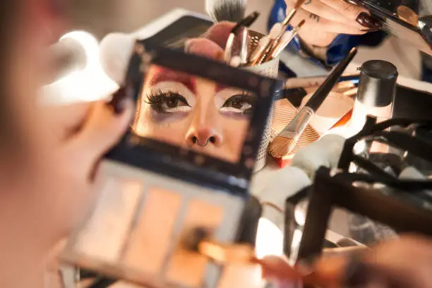 Reflexion at the hand mirror of the face of drag queen making bright makeup at the dressing table at home. Drag queen concept. Stock photo