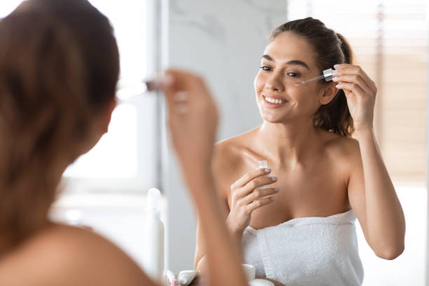 Woman Applying Face Serum Using Dropper Caring For Skin Indoors Facial Skincare. Young Woman Applying Face Serum Using Dropper Caring For Skin Standing Near Mirror In Modern Bathroom Indoors. Beauty Routine And Anti-Aging Skin Treatment Concept. Selective Focus woman wrapped in bath towel stock pictures, royalty-free photos & images