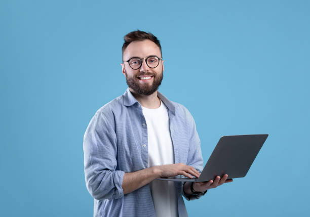 istock Cheerful millennial guy in glasses using laptop computer for online work or communication on blue studio background 1309247055