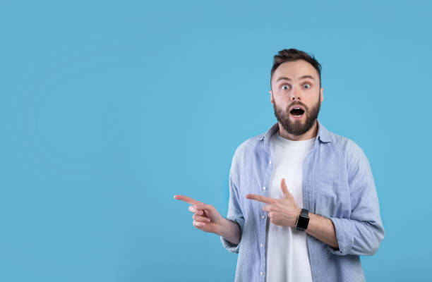 istock Shocked millennial man pointing at empty space in disbelief, standing with open mouth over blue background, free space 1309247479