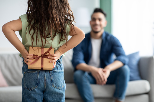 Little girl greeting her arabic dad with Fathers Day, holding gift behing back. Female child making surpsise to daddy at home, hiding present box, congratulating with holiday, selective focus