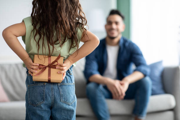Little girl greeting her dad with Fathers Day, holding gift behing back Little girl greeting her arabic dad with Fathers Day, holding gift behing back. Female child making surpsise to daddy at home, hiding present box, congratulating with holiday, selective focus fathers day gift stock pictures, royalty-free photos & images