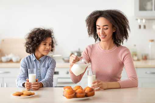 Time For Vitamins Concept. Smiling young African American woman pouring fresh milk from bottle into glass. Happy mother and little girl having delicious breakfast together in the kitchen