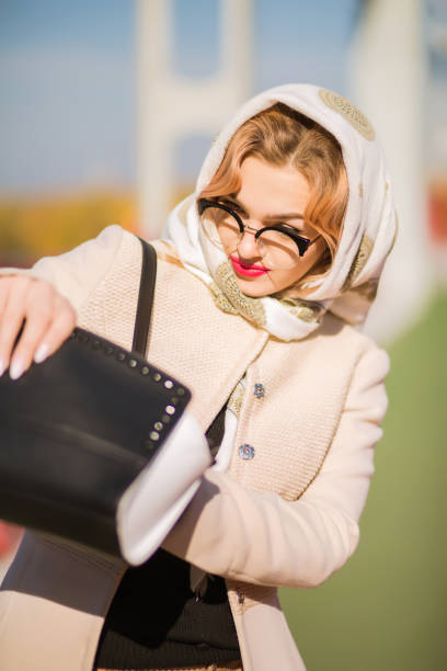 Stylish dressed woman looking into her handbag, wears beige coat and glasses