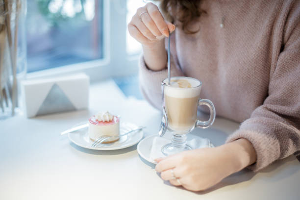 una mujer sosteniendo una cuchara larga en tu mano. taza de cristal con capuchino y pastel en un plato. - women spoon tasting elegance fotografías e imágenes de stock