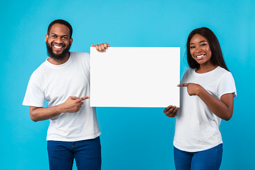 Cheerful Black Couple Holding Blank Paper Poster Together And Pointing At Free Space For Your Text Smiling To Camera Standing Over Blue Studio Background. People With White Board For Advertising