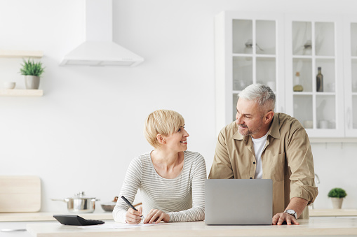 Smiling senior couple spouses use laptop, calculator, new technology paying banking bills online. Happy modern male and lady manage domestic finances expenditures considered family budget in kitchen
