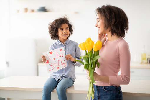 Happy Holiday. Portrait of cheerful black daughter making surprise for her mum and showing I Love You Mommy paper poster, greeting smiling woman with Mother's day or birthday sitting on table