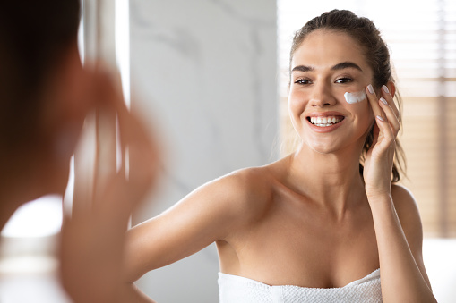 Smiling Woman Applying Face Cream Under Eyes Moisturizing Skin Posing Near Mirror Standing In Modern Bathroom At Home. Beauty Routine, Facial Skincare Concept. Selective Focus
