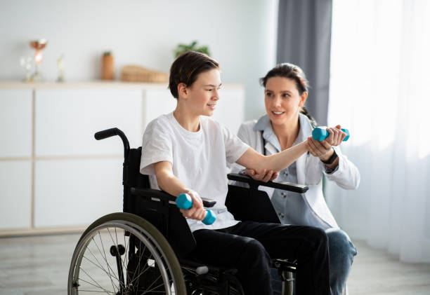 physikalisches rehabilitationskonzept. junger physiotherapeut hilft teenager im rollstuhl, übungen zu hause zu machen - lähmung stock-fotos und bilder