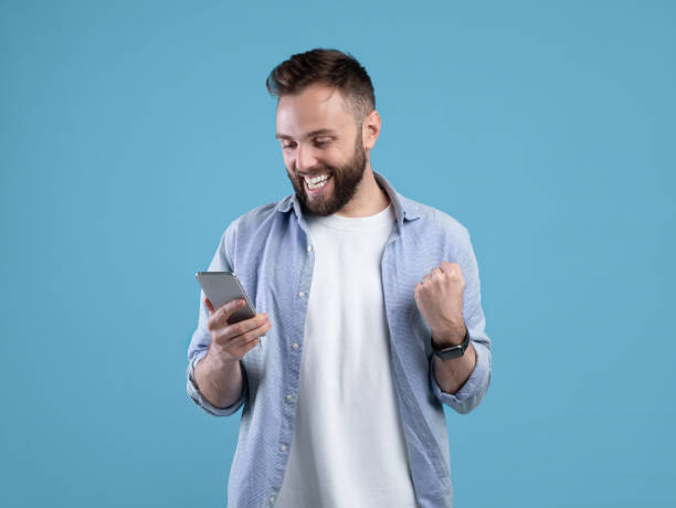istock Portrait of happy bearded young man using smartphone, making winner gesture over blue studio background 1309256870