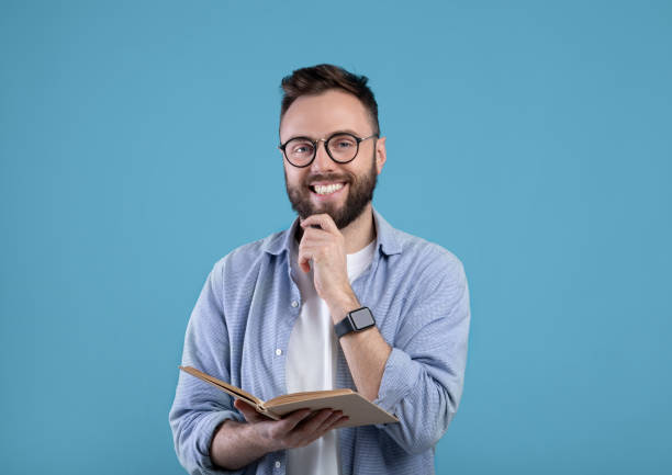 istock Happy young man with book looking at camera and smiling on blue studio background 1309256992
