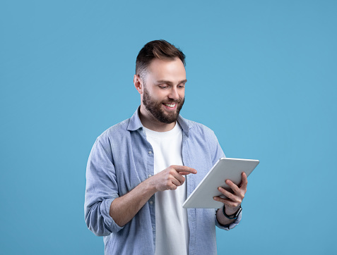 Joyful bearded man using tablet computer for online work or studies on blue studio background. Positive teacher giving remote lesson. Blogger creating new post for social network