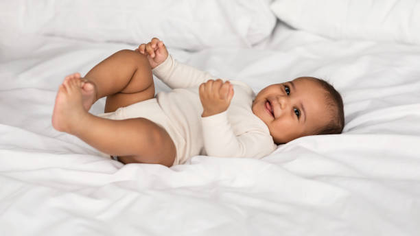 Cute little African American infant lying in bed In The Morning. Portrait of adorable smiling black infant lying on the back in bed on white bedsheet at home. Positive healthy laughing afro child wearing bodysuit, looking at camera. Happy Childhood cute baby laughing stock pictures, royalty-free photos & images