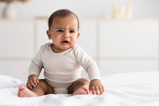 Portrait of sad upset African American baby crying, sitting on the bed in bedroom at home, wearing bodysuit. Unsatisfied hungry child whining, copy space, selective focus, blurred background