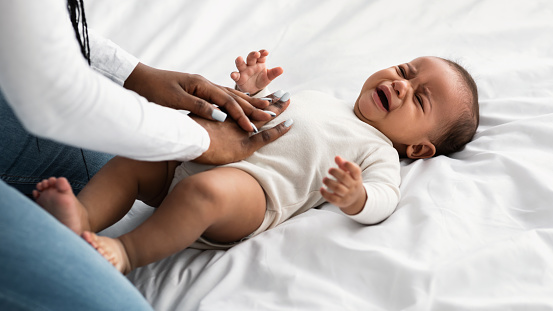 Displeased black baby crying and whining, feeling colic or abdomen ache, lying on back in bed. African American mother massaging her child's belly for pain relief. Parenthood And Maternity Concept