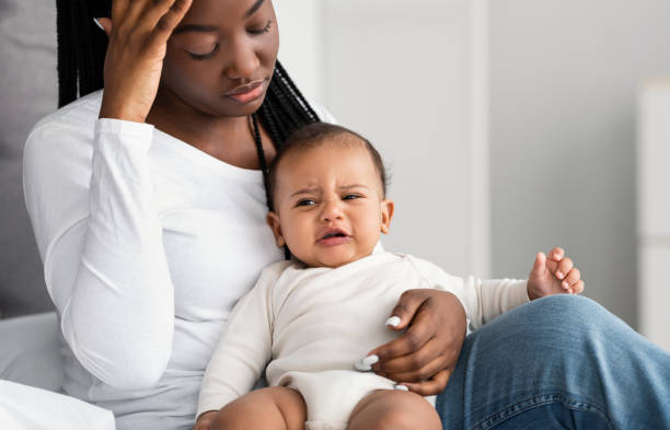 Stressed African American mom sitting with kid on bed Postnatal Stress Concept. Portrait of depressed African American woman sitting with her crying little child on bed at home, feeling ppd baby blues, headache or migraine, touching forehead postpartum depression stock pictures, royalty-free photos & images