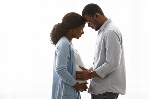 Sensual pregnant african american couple cuddling while spending time together at home, white background, copy space. Happy expecting black couple standing next to window and hugging woman big belly