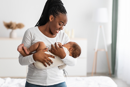 Maternity Concept. Portrait of smiling African American woman holding her adorable small black upset baby on hands, singing lullaby to sleep, comforting infant kid standing in bedroom at home