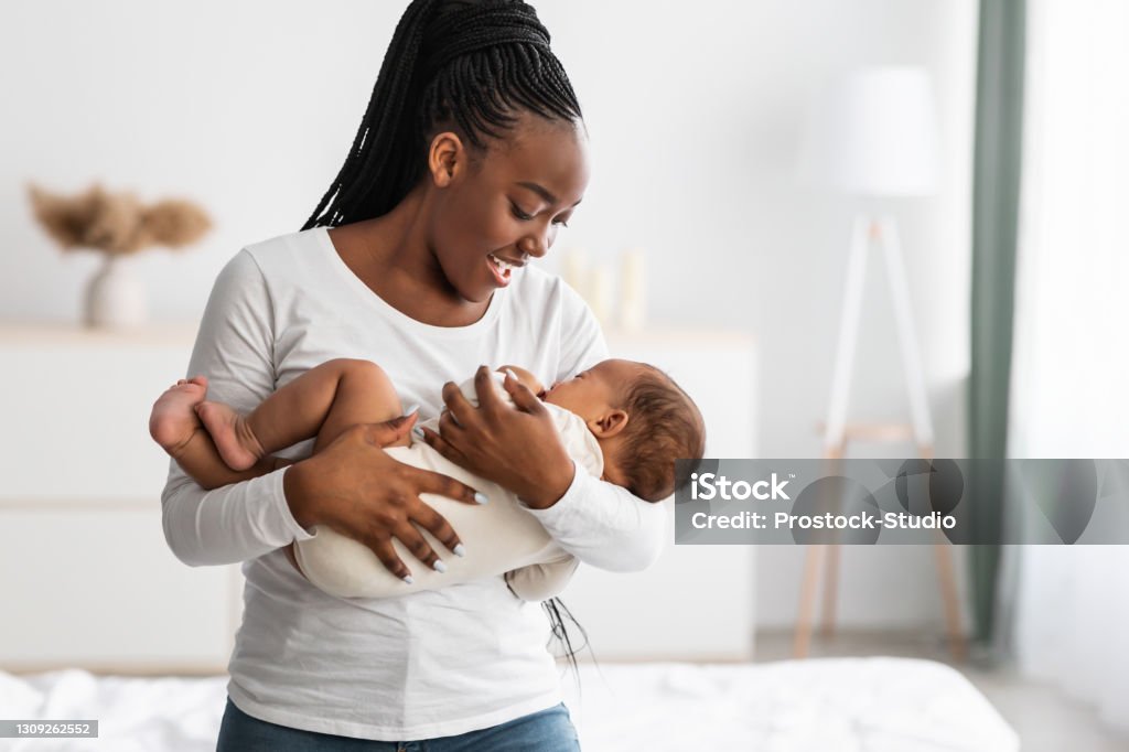 African American mother singing lullaby for infant to sleep Maternity Concept. Portrait of smiling African American woman holding her adorable small black upset baby on hands, singing lullaby to sleep, comforting infant kid standing in bedroom at home Breastfeeding Stock Photo