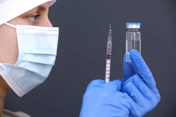 Doctor or nurse in medical mask and gloves is holding syringe and flu, measles, coronavirus vaccine in glass bottle for diseases outbreak vaccination, medicine and drug concept on gray background. el médico o enfermero con máscara médica y guantes está sosteniendo la vacuna contra la jeringa y la gripe, el sarampión, el coronavirus en el frasco de vidrio para la vacunación contra brotes de enfermedades, la medicina y el concepto de fármaco so