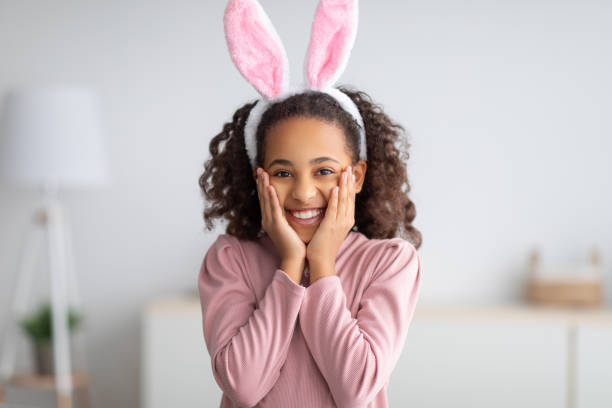 Positive black girl with rabbit ears smiling to camera and touching cheeks with hands
