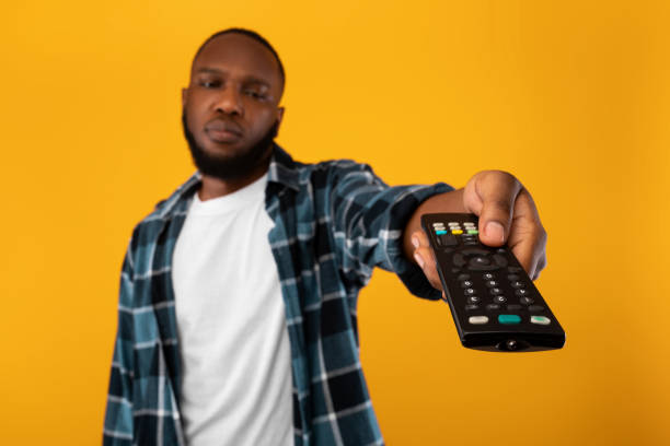 istock Bored African Man Switching Channels Pointing Television Controller, Yellow Background 1309263097