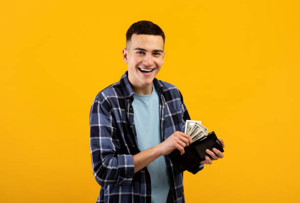 Smiling young man putting money into his wallet on orange studio background