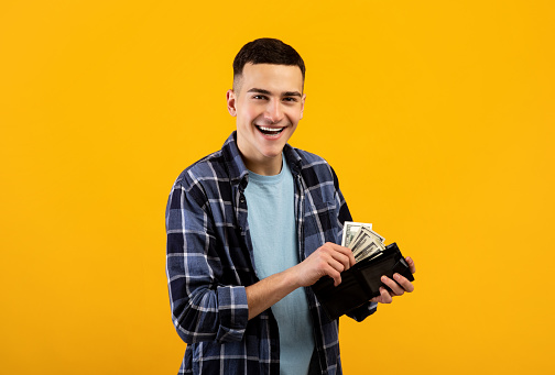 Smiling young man putting money into his wallet on orange studio background. Cool millennial guy holding purse full of dollars, being happy over his financial success or good income