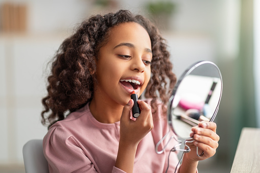 Daily make up. Happy teenage lady applying lipstick, holding mirror and looking at herself. Curly african american girl getting ready for school or date
