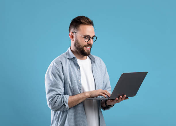 istock Confident young guy in glasses using laptop pc for online work or communication on blue studio background 1309265312