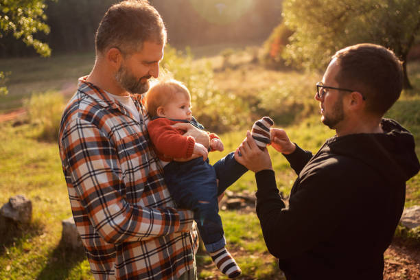 Gay couple with their baby boy in nature Gay couple with their child in nature, fixing baby socks to their son. gay daddy stock pictures, royalty-free photos & images