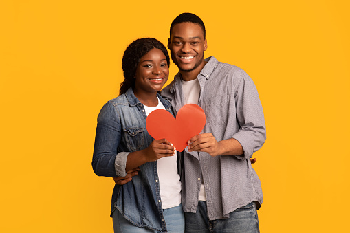 Joyful Loving Black Couple Holding Red Paper Heart And Hugging, Happy African American Man And Woman Celebrating Valentine's Day Together, Standing Isolated Over Yellow Background, Free Space