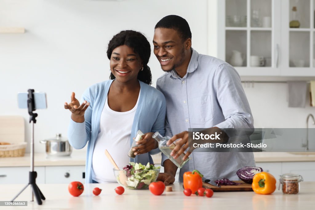 Happy black expecting family broadcasting while cooking Happy black expecting family broadcasting while cooking healthy food at home, using mobile phone on tripod. Cheerful african american pregnant couple having cooking lesson online, copy space Adult Stock Photo