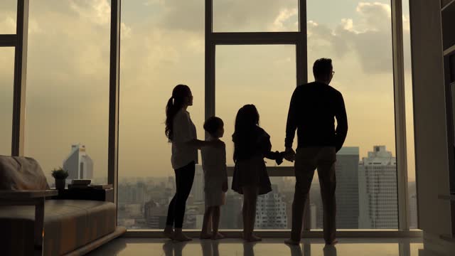 Silhouette rear view asian chinese family with 2 children looking through window enjoying beautiful sunset at living room in kuala lumpur city
