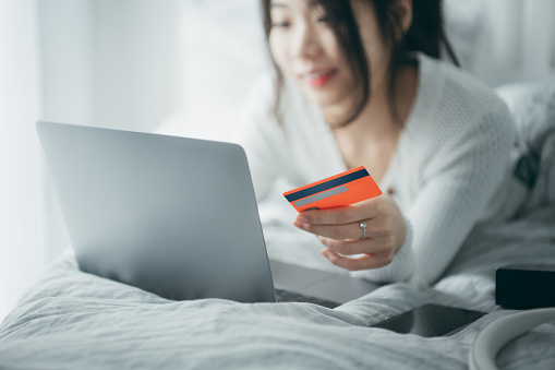 Asian young woman using her laptop while holding a credit card on bed at home.
