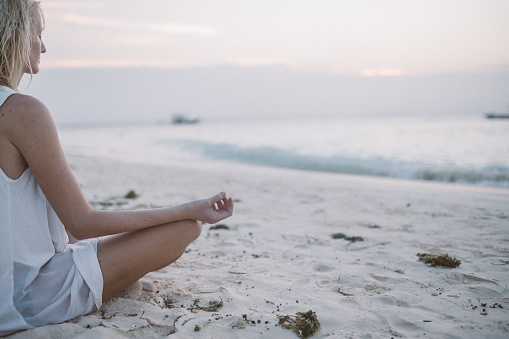 Young woman sitting on the beach and meditating. Balancing the energy in yoga style. Sea in background.