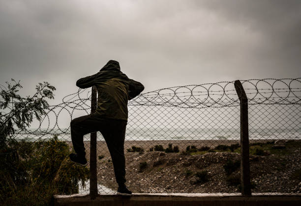 l'uomo rifugiato sta scalando una recinzione di rasoio di filo vicino alla spiaggia in una giornata nuvolosa - topics barbed wire fence chainlink fence foto e immagini stock
