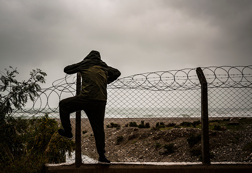 Refugee man is climbing a wire razor fence near the beach on a overcast day