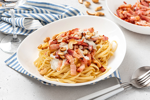 Pasta with creamy sauce, fried bacon and nuts in a white plate on a light background close-up. Pasta with bolognese and ham.