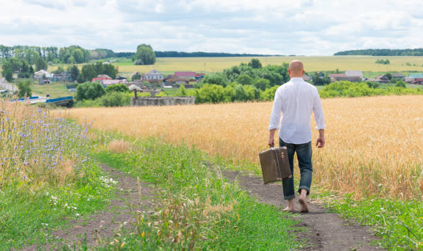 A person returns after a trip to their homeland. A brutal young man in a white shirt is walking barefoot on the way home, holding an old large suitcase. A person returns after a trip to their homeland. The wanderer is on his way home. tramp home stock pictures, royalty-free photos & images