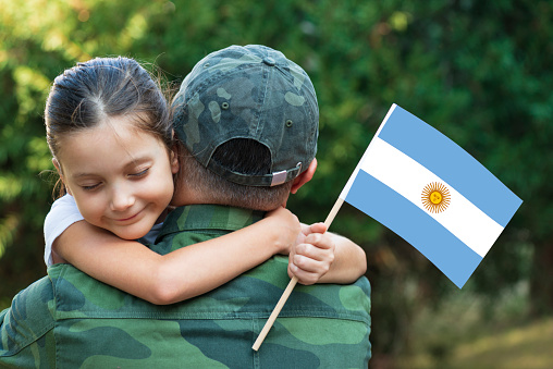 Little girl hugs the soldier father and holding Argentinian flag.