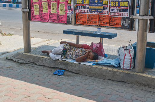 Kolkata, 03-25-2021: A poor aged woman sleeping on footpath, under a bus shelter. She has a bottle of water and some luggage beside her. Photo taken during afternoon in a hot summer day.