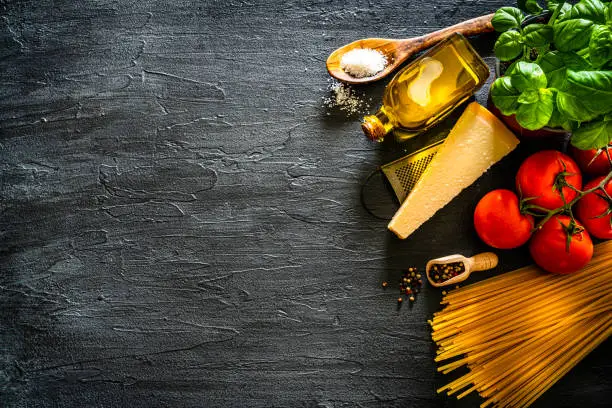 Italian food background: overhead view of a black table with uncooked spaghetti and ingredients for cooking a traditional Italian pasta arranged at the right of a black background making a frame and leaving useful copy space. The composition includes basil. fresh ripe tomatoes, olive oil, garlic, pepper, salt and Parmesan cheese. High resolution 42Mp studio digital capture taken with Sony A7rII and Sony FE 90mm f2.8 macro G OSS lens
