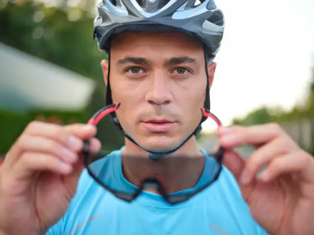 Photo of Portrait of handsome young male cyclist looking at camera while holding, putting on protective glasses, getting ready for cycling outdoors