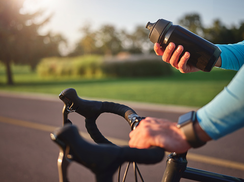 Hydrate your body. Close up of hands of professional cyclist holding water bottle, standing with his bike in park on a sunny day. Sport and recreation concept. Selective focus