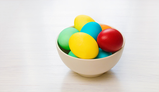 Multicolored easter eggs in bowl close up. White bowl with painted easter eggs on wooden table, side view. Template for design to easter holiday