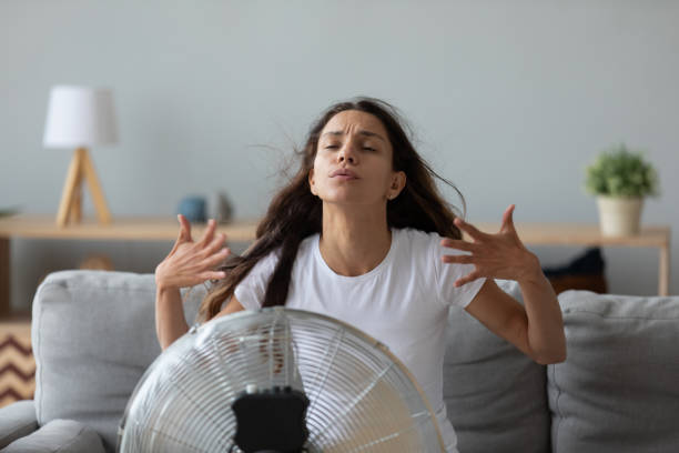 Funny overheated woman enjoying fresh air, cooling by electric fan Funny overheated woman enjoying fresh air, cooling by electric fan, exhausted young female waving hands, sitting on couch at home in front of ventilator, suffering from hot summer weather hyperthermia photos stock pictures, royalty-free photos & images